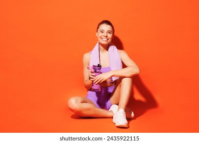Full body smiling young fitness trainer instructor woman sportsman wear top shorts purple clothes in home gym sit drink water relax rest isolated on plain orange background. Workout sport fit concept