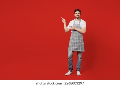 Full body smiling young male housewife housekeeper chef cook baker man wear grey apron point index finger aside on workspace area mock up isolated on plain red background studio. Cooking food concept - Powered by Shutterstock