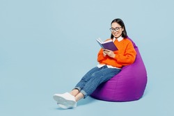 Full Body Smiling Young Woman Of Asian Ethnicity Wear Orange Sweater Glasses Sit In Bag Chair Read Book At Library Isolated On Plain Pastel Light Blue Cyan Background Studio. People Lifestyle Concept