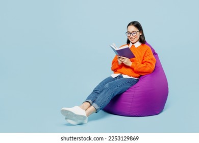 Full body smiling young woman of Asian ethnicity wear orange sweater glasses sit in bag chair read book at library isolated on plain pastel light blue cyan background studio. People lifestyle concept - Shutterstock ID 2253129869