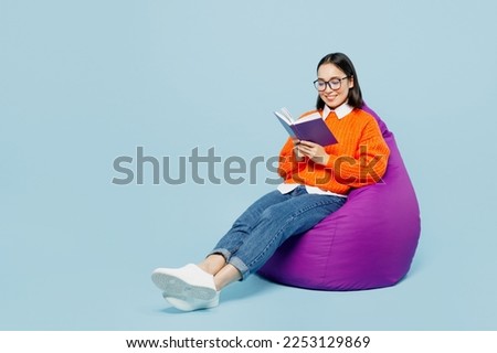 Full body smiling young woman of Asian ethnicity wear orange sweater glasses sit in bag chair read book at library isolated on plain pastel light blue cyan background studio. People lifestyle concept
