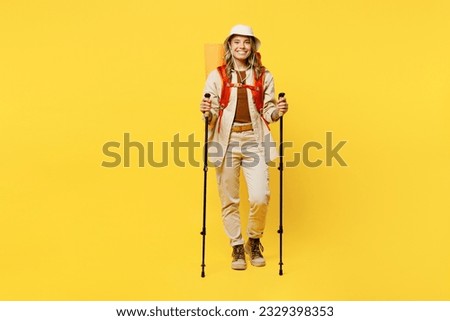 Full body smiling young woman carry bag with stuff mat hold trekking poles isolated on plain yellow background. Tourist leads active lifestyle walk on spare time. Hiking trek rest travel trip concept