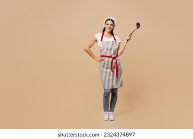 Full body smilng happy fun young housewife housekeeper chef baker latin woman wear apron toque hat hold in hand laddle look camera isolated on plain pastel light beige background. Cook food concept