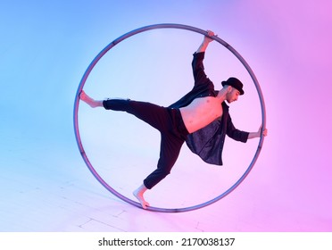 Full body of sportive male acrobat in hat performing waltz stunt with cyr wheel during training against light background in studio