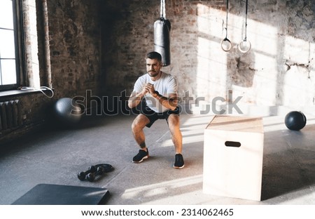 Full body of strong male athlete doing squats while training muscles stretching legs standing in chair pose during workout at gym