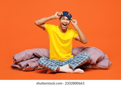 Full body surprised happy young man wear pyjamas jam take off sleep eye mask sit with duvet blanket rest relax at home isolated on plain orange background studio portrait. Good mood night nap concept