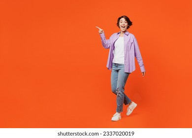 Full body surprised shocked ecxited young woman she wears purple shirt white t-shirt casual clothes walk go point finger aside isolated on plain orange background studio portrait. Lifestyle concept - Powered by Shutterstock