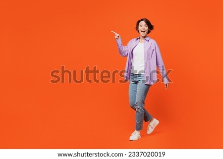 Full body surprised shocked ecxited young woman she wears purple shirt white t-shirt casual clothes walk go point finger aside isolated on plain orange background studio portrait. Lifestyle concept