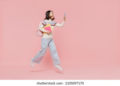 Full body teen student girl of Asian ethnicity in sweater hold backpack jump high use mobile cell phone jump high isolated on pastel plain light pink background Education in university college concept