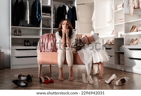 Full body of thoughtful young female sitting on sofa and pouting lips while picking out high heeled shoes in living room near white closet at home