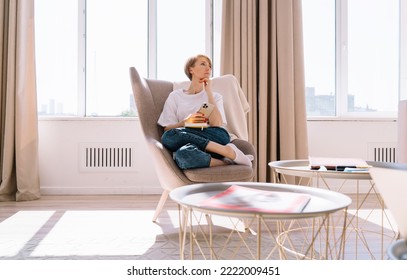 Full body of thoughtful young woman with short hair sitting in gray armchair with hand at chin and using cellphone while resting in modern living room at home during weekend - Powered by Shutterstock