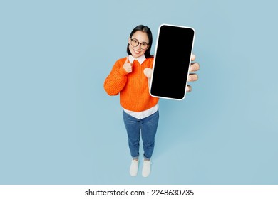 Full body top view from above young woman of Asian ethnicity wear orange sweater glasses hold in hand use mobile cell phone with blank screen area isolated on plain pastel light blue cyan background