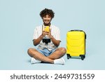 Full body traveler Indian man wear white casual clothes sit near bag use mobile cell phone isolated on plain blue background. Tourist travel abroad in free time rest getaway. Air flight trip concept