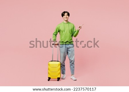 Full body traveler man of Asian ethnicity wear green hoody hold suitcase valise isolated on plain pastel pink background. Tourist travel abroad in free spare time rest. Air flight trip journey concept