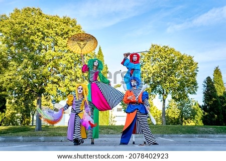 Full body troupe of funny talented performers with painted faces in colorful clown costumes and wigs juggling pins and performing dance on stilts during parade, in city park