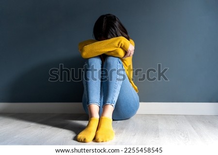 Full body of unrecognizable unhappy young lady with dark hair in casual clothes embracing knee and covering face while sitting on floor near wall