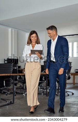 Full body vertical portrait of walking mature Latin businessman and European businesswoman discussing project on tablet in office. Two diverse partners of professional business people work together.