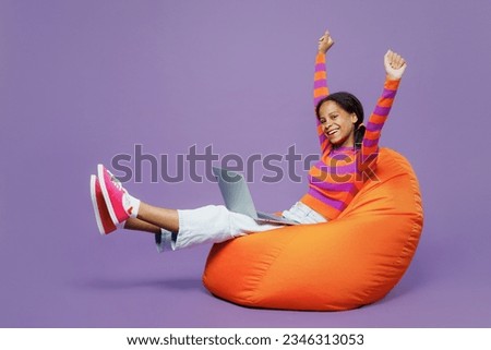Full body winner little kid teen IT girl 15-16 years old wear striped orange sweatshirt sit in bag chair hold use laptop pc computer isolated on plain purple background. Childhood lifestyle concept