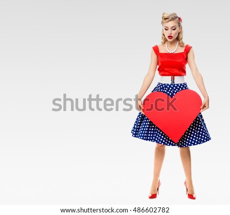Full body of woman with heart symbol, pin-up style dress with polka dot, on grey, blank copyspace area for text, slogan. Caucasian blond model posing in retro fashion and vintage concept studio shoot.