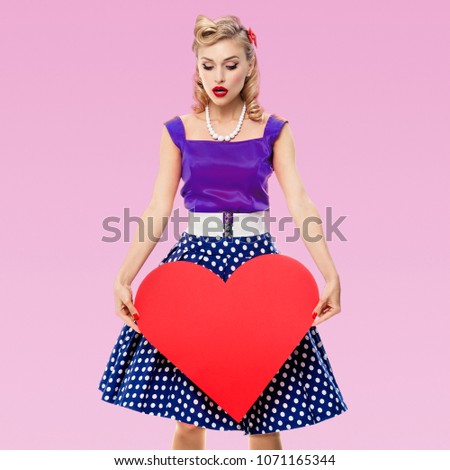 Full body of woman holding heart symbol, dressed in pin-up style dress with polka dot, on pink background. Caucasian blond model posing in retro fashion and vintage concept studio shoot.