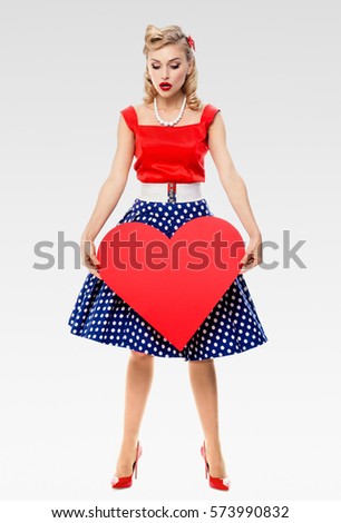 Full body of woman holding heart symbol, dressed in pin-up style dress with polka dot, on grey background. Caucasian blond model posing in retro fashion and vintage concept studio shoot.