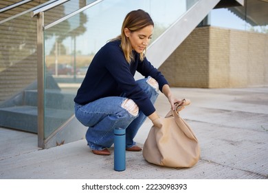 Full Body Woman In Stylish Clothes Sitting On Haunches Near Steps And Searching For Keys Inside Bag Outside Modern Apartment Building