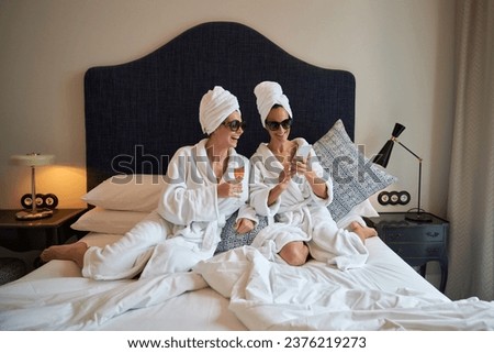 Full body of woman in sunglasses and terry bathrobe and towel turban sitting on bed with glass of champagne and looking at female friend browsing smartphone