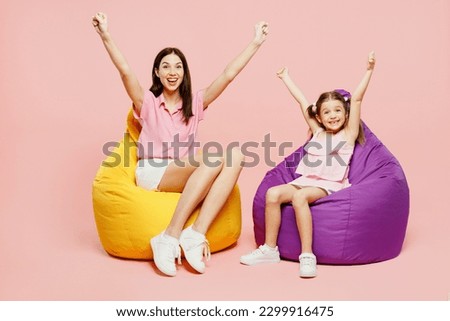 Full body woman wear casual clothes with child kid girl 6-7 years old. Mother daughter sit in bag chair do winer gesture look camera isolated on plain pastel pink background. Family parent day concept