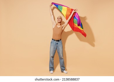 Full body young activist bisexual happy fun blond latin gay man with make up in beige tank shirt waving hold rainbow flag isolated on plain light ocher background studio People lgbt lifestyle concept.