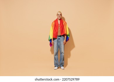 Full Body Young Activist Smiling Happy Fun Cool Blond Latin Gay Man With Make Up In Beige Tank Shirt Wrapped Rainbow Flag Isolated On Plain Light Ocher Background Studio People Lgbt Lifestyle Concept.