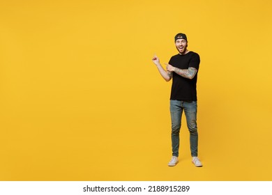 Full Body Young Amazed Bearded Tattooed Man 20s He Wears Casual Black T-shirt Cap Indicate Point Index Fingers Aside On Workspace Area Mock Up Isolated On Plain Yellow Wall Background Studio Portrait.