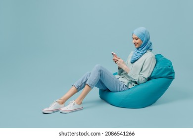 Full body young arabian asian muslim woman in abaya hijab sit in bag chair hold in hand use mobile cell phone isolated on plain blue background studio People uae middle eastern islam religious concept