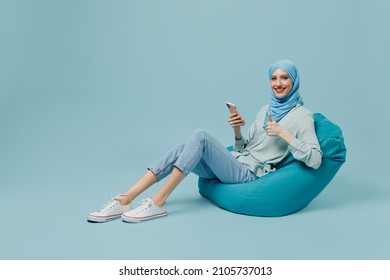 Full body young arabian asian muslim woman in abaya hijab sit in bag chair hold use mobile cell phone show thumb up isolated on plain blue background People uae middle eastern islam religious concept
