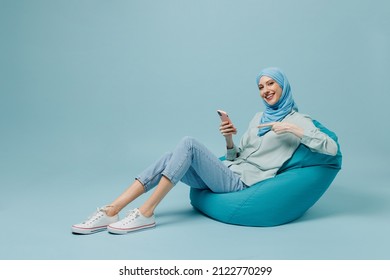 Full body young arabian asian muslim woman in abaya hijab sit in bag chair hold point on mobile cell phone isolated on plain blue background studio People uae middle eastern islam religious concept