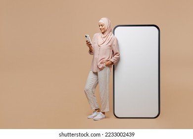 Full body young arabian asian muslim woman she in abaya hijab clothes big huge blank screen mobile cell phone use smartphone isolated on plain light beige background People uae islam religious concept - Shutterstock ID 2233002439