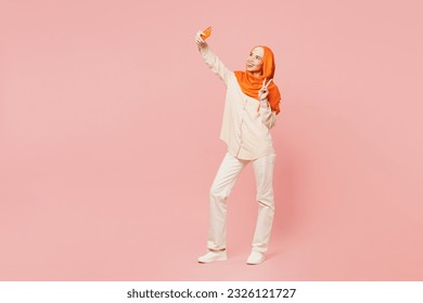 Full body young arabian asian muslim woman wearing orange abaya hijab doing selfie shot on mobile cell phone show v-sign isolated on plain pink background. Uae middle eastern islam religious concept - Powered by Shutterstock