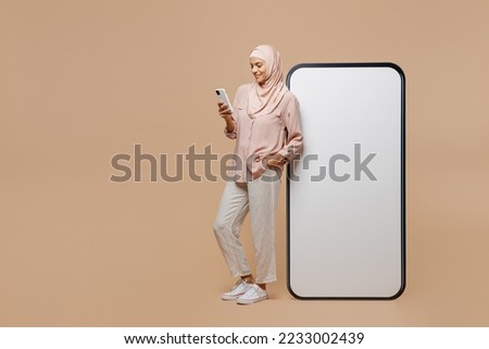 Full body young arabian asian muslim woman she in abaya hijab clothes big huge blank screen mobile cell phone use smartphone isolated on plain light beige background People uae islam religious concept