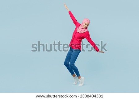 Full body young arabian asian muslim woman wear pink abaya hijab stand on toes leaning back with outstretched hands dance isolated on plain pastel light blue cyan background. People uae islam concept