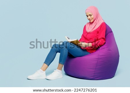 Full body young arabian muslim woman wear pink abaya hijab sit in bag chair read book novel diary isolated on plain pastel light blue cyan background studio portrait People uae islam religious concept