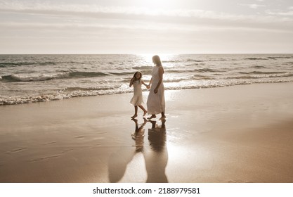 Full body of young barefooted woman with long hair in maxi white dress holding hand of adorable little daughter, while walking together along wet sandy beach near wavy ocean - Powered by Shutterstock