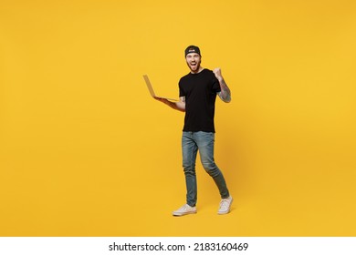 Full Body Young Bearded Tattooed Man 20s He Wears Casual Black T-shirt Cap Hold Use Work On Laptop Pc Computer Do Winner Gesture Isolated On Plain Yellow Wall Background Studio. Tattoo Translate Fun