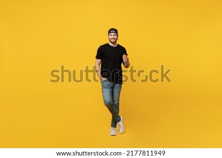 Full body young bearded tattooed man 20s he wears casual black t-shirt cap hold takeaway delivery craft paper brown cup coffee to go isolated on plain yellow wall background. People lifestyle concept