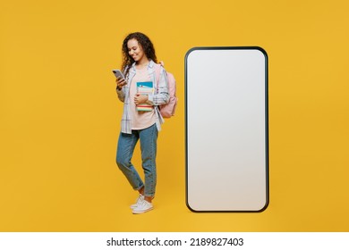 Full Body Young Black Teen Girl Student Wear Casual Clothes Backpack Bag Hold Books Big Huge Blank Screen Use Mobile Cell Phone Mockup Isolated On Plain Yellow Background. High School College Concept