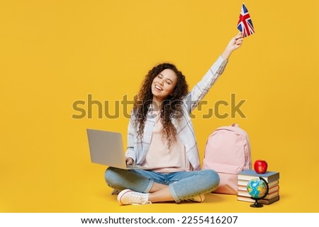 Full body young black teen girl student wear casual clothes backpack bag use work on laptop pc computer raise up british flag isolated on plain yellow background High school university college concept
