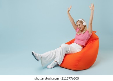 Full body young blonde woman 20s in casual pink t-shirt sit in bag chair headohones listen to music audiobook stretch hands isolated on plain pastel light blue background. People lifestyle concept.