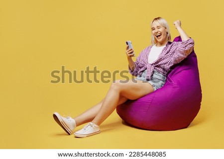 Full body young blonde woman 20s she in pink tied shirt white t-shirt sit in bag chair hold in hand use mobile cell phone do winer gesture isolated on plain yellow background People lifestyle concept