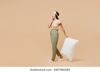 Full body young calm Latin woman she wears pyjamas jam sleep eye mask rest relax at home walk go hold pillow cover mouth with hand yawn isolated on plain beige background. Good mood night nap concept