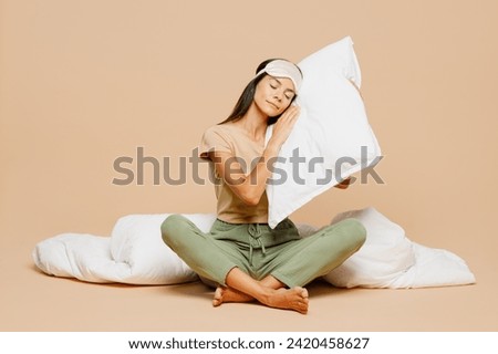 Full body young calm Latin woman wears pyjamas jam sleep eye mask rest relax at home sitting put head on pillow hold duvet close eyes isolated on plain beige background. Good mood night nap concept