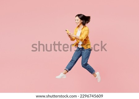 Full body young cheerful woman of Asian ethnicity wear yellow shirt white t-shirt jump high hold in hand use mobile cell phone run fast isolated on plain pastel light pink background studio portrait
