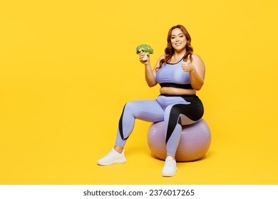 Full body young chubby plus size big fat fit woman wear blue top warm up training sit on fit ball eat broccoli show thumb up isolated on plain yellow background studio home gym. Workout sport concept